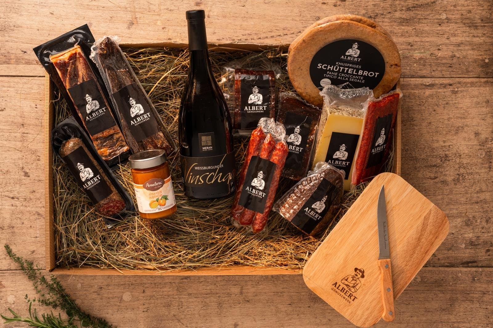 Holzkiste mit Rotwein, Speck, Kaminwurzen, Käse, Marmelade, Schüttelbrot sowie Speckbrettl und Messer auf Stroh