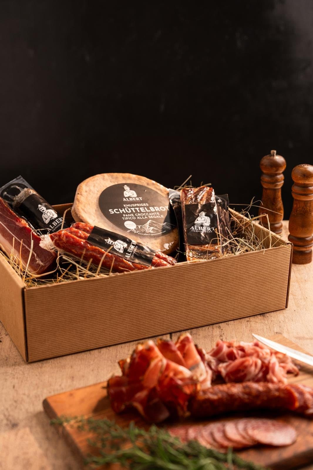 Südtiroler Geschenkbox mit Schüttelbrot, Speck und Kaminwurzen auf Stroh, davor ein Brett mit aufgeschnittenem Speck