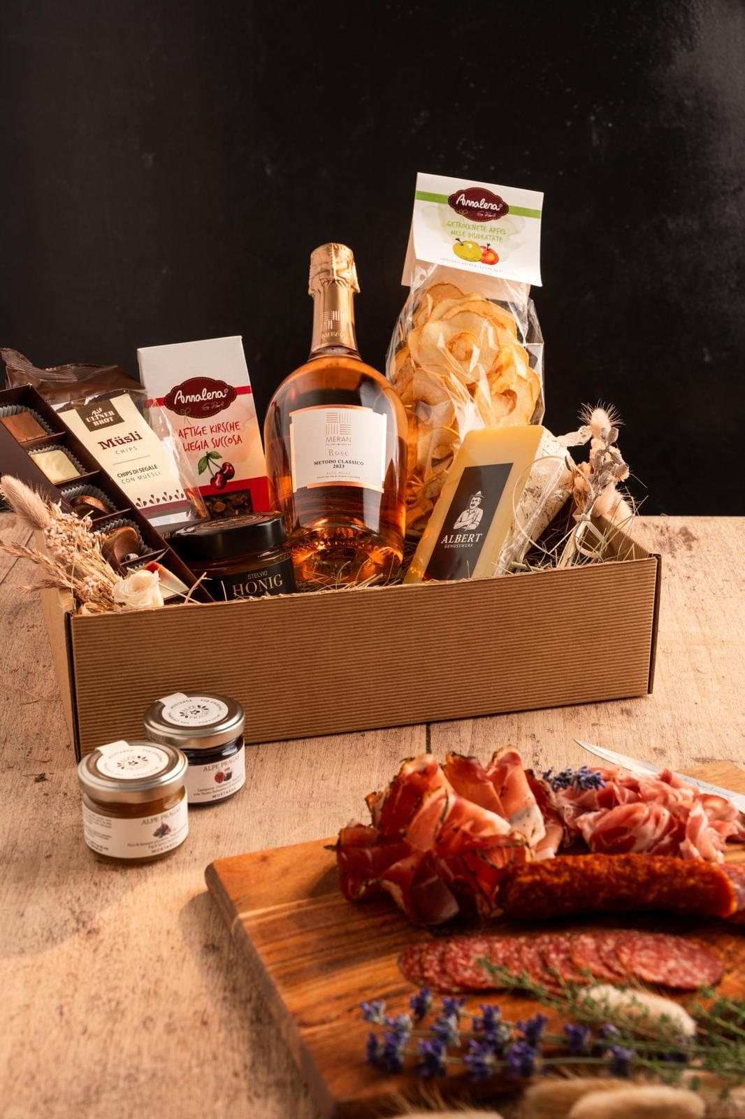 Geschenkbox mit Prosecco, Nudeln, Aufstrichen und Käse, davor ein Brett mit Aufschnitt