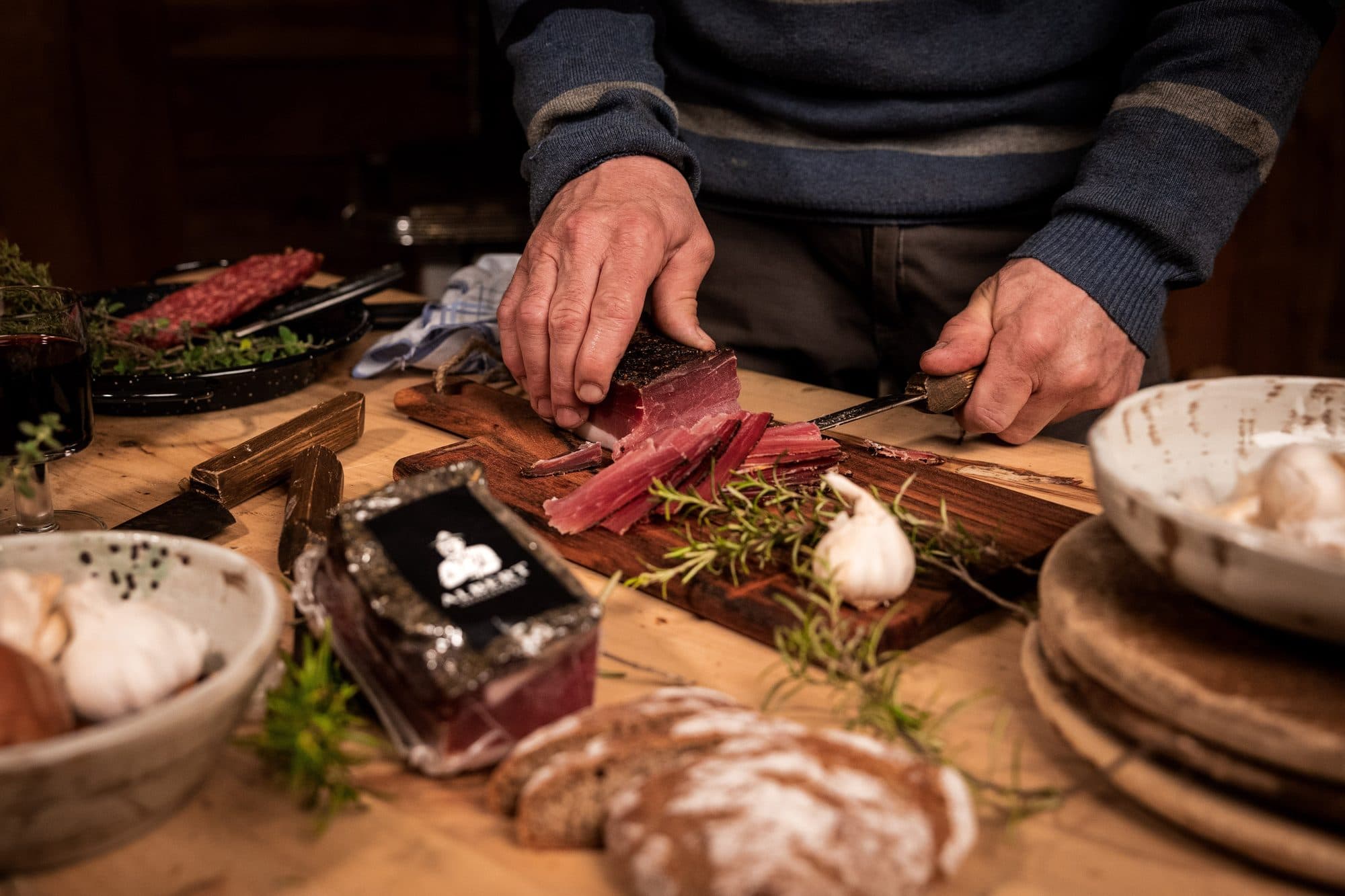 Hände schneiden Südtiroler Speck auf einem Holzbrett, daneben Brot, Knoblauch und Rosmarin